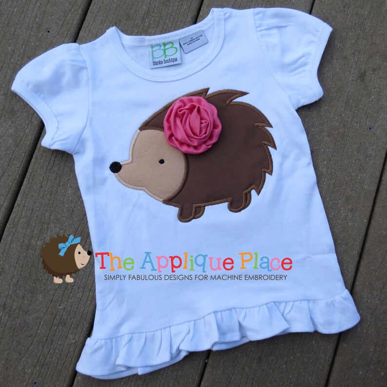 Puede incluir: Una camiseta azul claro con una aplicaci&oacute;n de erizo marr&oacute;n con una flor rosa en la parte superior. La camiseta tiene un dobladillo con volantes y el texto "The Applique Place Simply Fabulous Designs for Machine Embroidery" est&aacute; impreso en la parte inferior.
