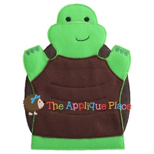 May include: A green and brown felt turtle puppet with a smiling face and outstretched arms. The puppet has a brown shell and the text "The Applique Place" in colorful letters.