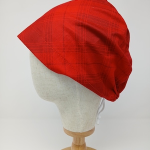 Puede incluir: Un sombrero rojo con estampado de cuadros, exhibido en una cabeza de maniquí. El sombrero es de un tono rojo vibrante con un diseño de cuadros rojos más oscuros. El sombrero tiene un cierre de cordón. La cabeza del maniquí está sobre un soporte de madera.