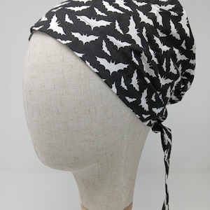 May include: A black and white patterned head covering featuring a repeating bat design. The fabric is tied at the back, and the head covering is displayed on a mannequin head. The head covering is likely for medical or culinary use.
