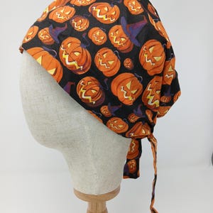 Puede incluir: Un gorro temático de Halloween con un patrón repetido de calabazas naranjas y sombreros de bruja morados sobre un fondo negro. El gorro está atado en la parte posterior con lazos de tela a juego. La tela está envuelta alrededor de una cabeza de maniquí.