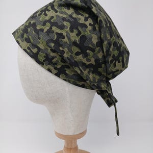 Könnte beinhalten: Ein Camouflage-Bandana in Grün-, Schwarz- und Grautönen, hinten gebunden. Das Bandana wird auf einem Schaufensterpuppenkopf mit Holzständer präsentiert. Der Stoff hat ein sich wiederholendes Muster aus unregelmäßigen Formen.