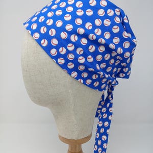 Könnte beinhalten: Blauer Baseball-Hut mit einem sich wiederholenden Muster aus weißen Basebällen mit roten Nähten. Der Hut ist hinten mit Stoffbändern gebunden. Präsentiert auf einem Schaufensterkopf.