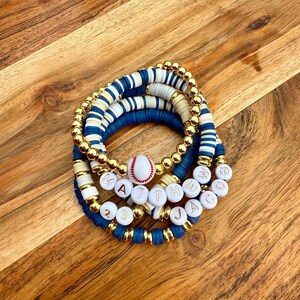May include: A set of four bracelets with white and blue beads, gold beads, and white beads with gold letters spelling out the name "MATTHEW JACOB" and the numbers "23". A white baseball with red stitching is included in the center of the bracelets.