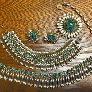 May include: A gold-tone jewelry set consisting of a necklace, a bracelet, a brooch, and a pair of clip-on earrings. Each piece features a textured, fan-like metal design accented with small, round, iridescent green and blue rhinestones.