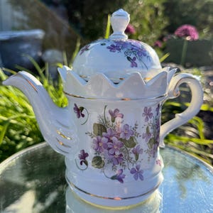 May include: A white porcelain teapot featuring a delicate purple violet floral pattern and gold trim. The teapot has a scalloped rim, a curved handle, a pointed spout, and a matching domed lid with a small knob handle on top.