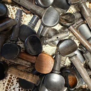 May include: A collection of vintage violin tuning pegs in various shades of brown and black. The pegs have rounded heads and tapered shafts, showing signs of age and wear. Some pegs have a dark, polished finish, while others are lighter and more weathered.
