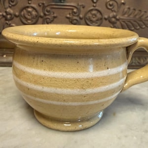 May include: A ceramic mug with a handle, featuring a light yellow glaze and horizontal white stripes. The mug has a rounded base and a wide rim. The mug is approximately 4 inches in diameter and 3 inches tall.