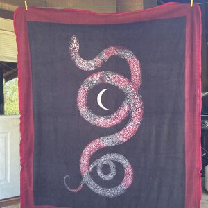 May include: A black fleece blanket with a red border features a white and red snake design with a crescent moon in the center.