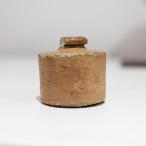 Puede incluir: Un pequeño tapón de botella marrón antiguo, cilíndrico, con una parte superior redondeada. El tapón está hecho de un material rugoso y texturizado y se encuentra sobre una superficie blanca. Una concha decorativa está en el fondo.