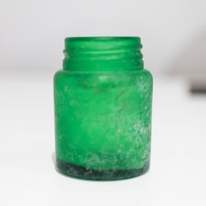 Könnte beinhalten: Kleine grüne Glasflasche mit strukturierter Oberfläche. Die Flasche hat eine kurze, zylindrische Form mit einem Gewindehals. Die Flasche ist leer und steht auf einer weißen Oberfläche.