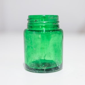 Pode incluir: Um pequeno frasco de vidro verde transparente com gargalo roscado. O frasco está vazio e assenta numa superfície branca. A cor verde é vibrante e o vidro tem um aspeto ligeiramente texturizado. O frasco é provavelmente para guardar pequenos objetos.