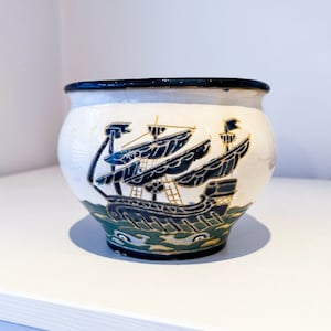 May include: A white ceramic pot with a blue and gold design of a ship with sails. The pot has a dark blue rim.