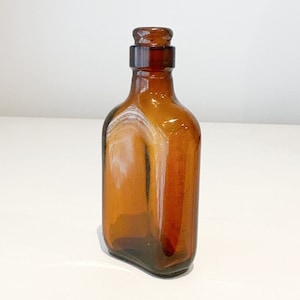 May include: A small, amber-colored glass bottle with a glass stopper. The bottle is empty and has a rectangular shape.