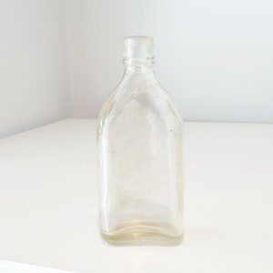 May include: A clear glass bottle with a rounded bottom and a square top. The bottle is empty and has a slightly textured surface.