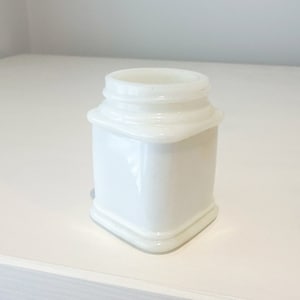 Pode incluir: Um pequeno frasco de vidro branco quadrado com um acabamento liso e brilhante. O frasco tem um design simples e elegante e é perfeito para guardar pequenos objetos.