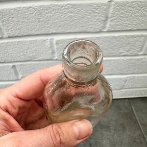 May include: A clear glass bottle with a rounded bottom and a slightly flared opening. The bottle has a textured surface and appears to be old.