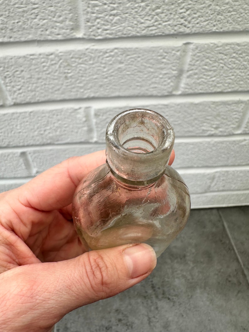 May include: A clear glass bottle with a rounded bottom and a slightly flared opening. The bottle has a textured surface and appears to be old.