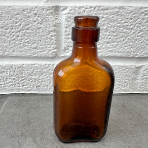 May include: A dark brown glass bottle with a rounded rectangular shape and a glass stopper. The bottle is sitting on a gray surface.