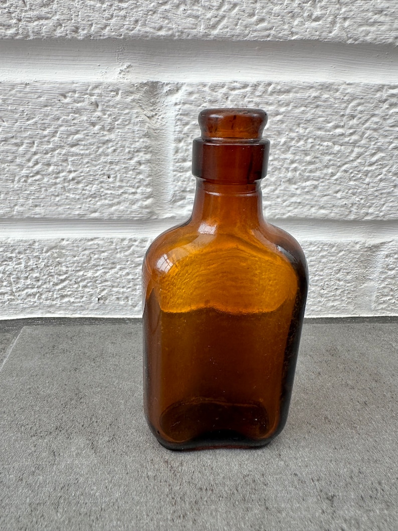 May include: A dark brown glass bottle with a rounded rectangular shape and a glass stopper. The bottle is sitting on a gray surface.