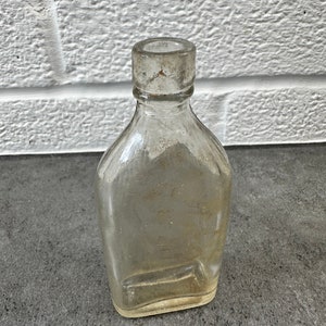 May include: A clear glass bottle with a rounded bottom and a short, wide neck. The bottle is empty and has a slightly cloudy appearance.