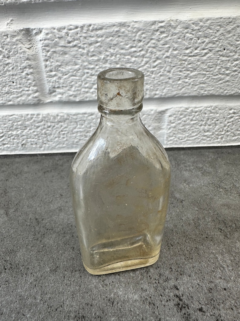 May include: A clear glass bottle with a rounded bottom and a short, wide neck. The bottle is empty and has a slightly cloudy appearance.