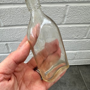 May include: A clear glass bottle with a rounded bottom and a narrow neck. The bottle is empty and has a slightly cloudy appearance.