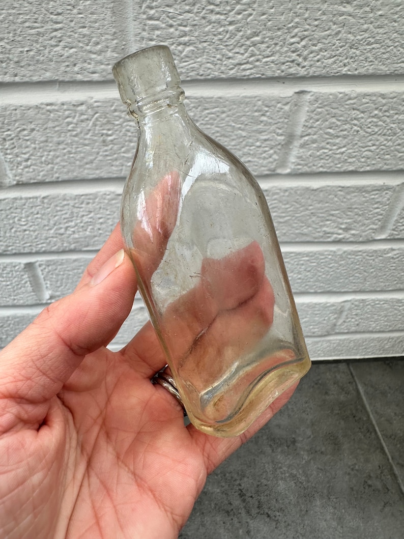 May include: A clear glass bottle with a rounded bottom and a narrow neck. The bottle is empty and has a slightly cloudy appearance.