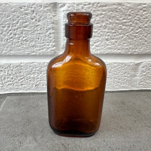 May include: A small, amber-colored glass bottle with a rounded, rectangular shape and a glass stopper. The bottle is sitting on a gray surface.