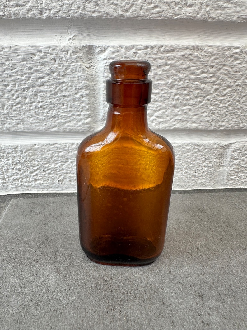 May include: A small, amber-colored glass bottle with a rounded, rectangular shape and a glass stopper. The bottle is sitting on a gray surface.