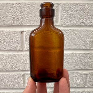 May include: A small, amber-colored glass bottle with a cork stopper. The bottle is held in a hand against a white brick wall background.