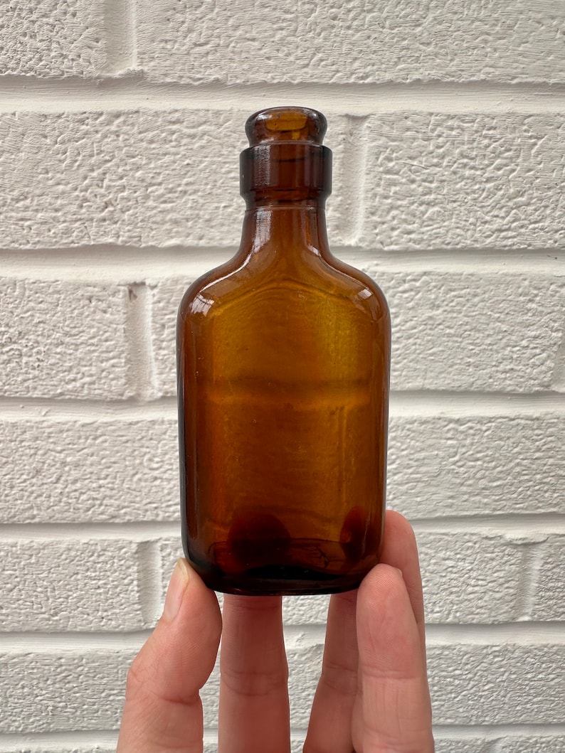 May include: A small, amber-colored glass bottle with a cork stopper. The bottle is held in a hand against a white brick wall background.