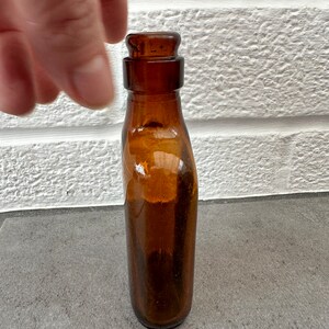 May include: A small, amber-colored glass bottle with a rounded top and a slightly flared bottom.