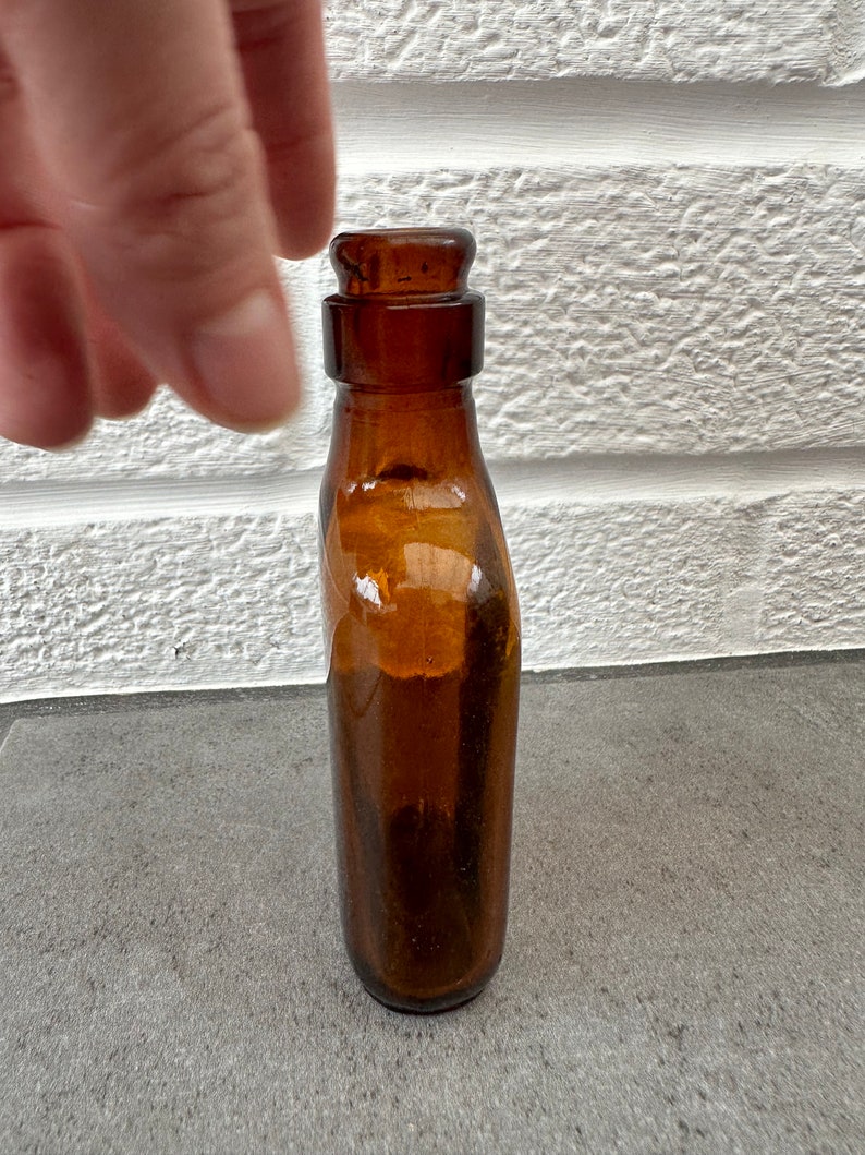 May include: A small, amber-colored glass bottle with a rounded top and a slightly flared bottom.