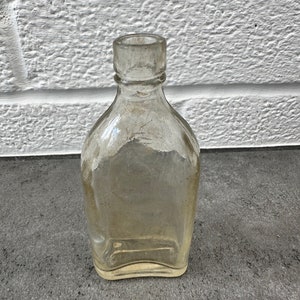 May include: A clear glass bottle with a rounded bottom and a square top. The bottle is empty and has a slightly cloudy appearance.