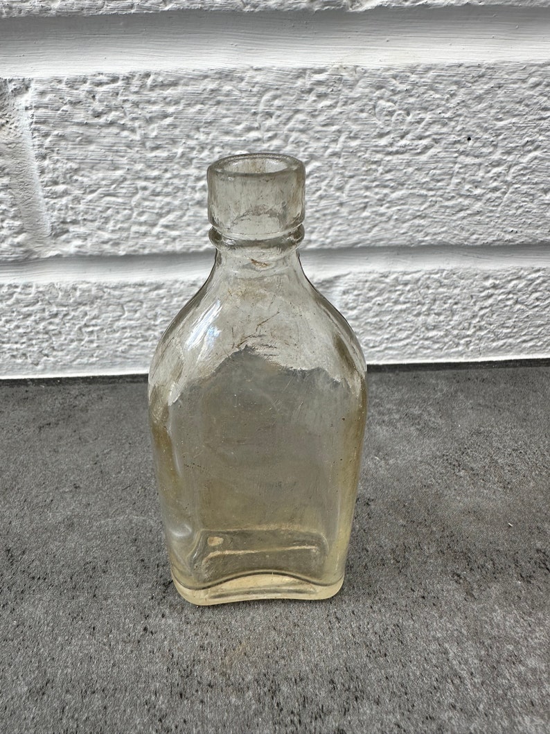 May include: A clear glass bottle with a rounded bottom and a square top. The bottle is empty and has a slightly cloudy appearance.
