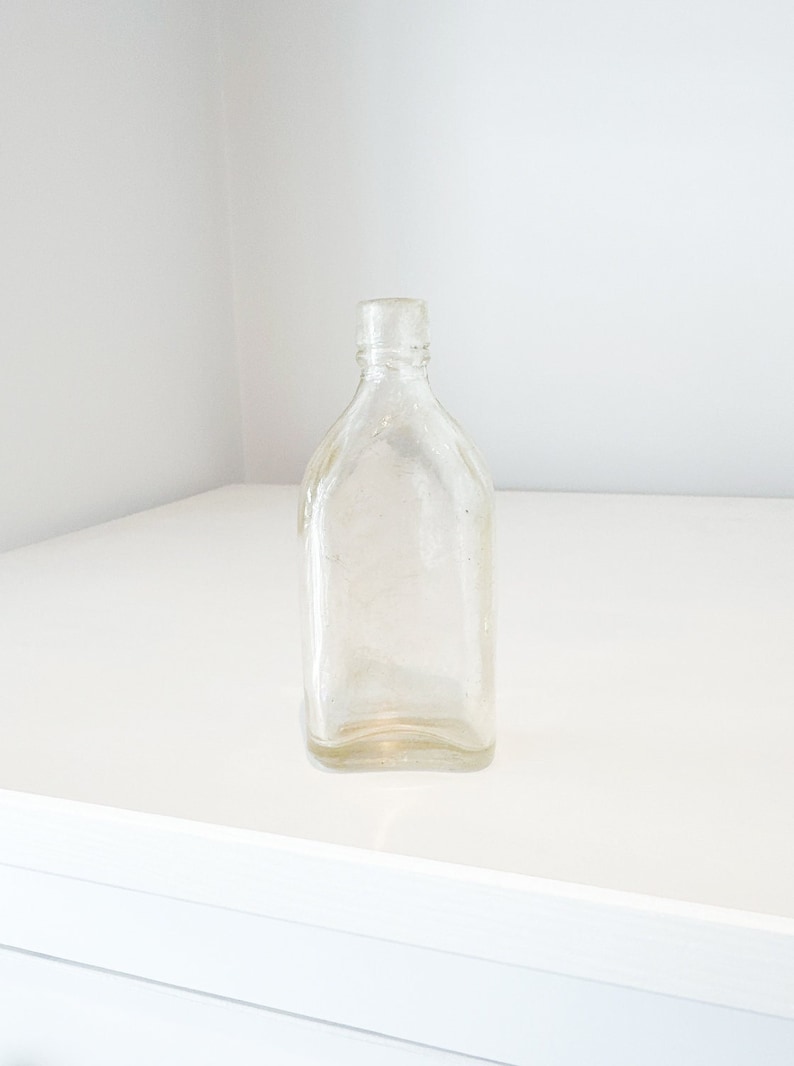 May include: A clear glass bottle with a rounded bottom and a square top. The bottle is empty and has a slightly textured surface.