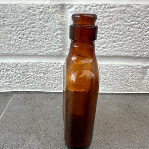 May include: A brown glass bottle with a rounded bottom and a short neck. The bottle has a glass stopper and is empty.