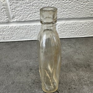 May include: A clear glass bottle with a rounded bottom and a slightly flared top. The bottle is empty and has a textured surface.