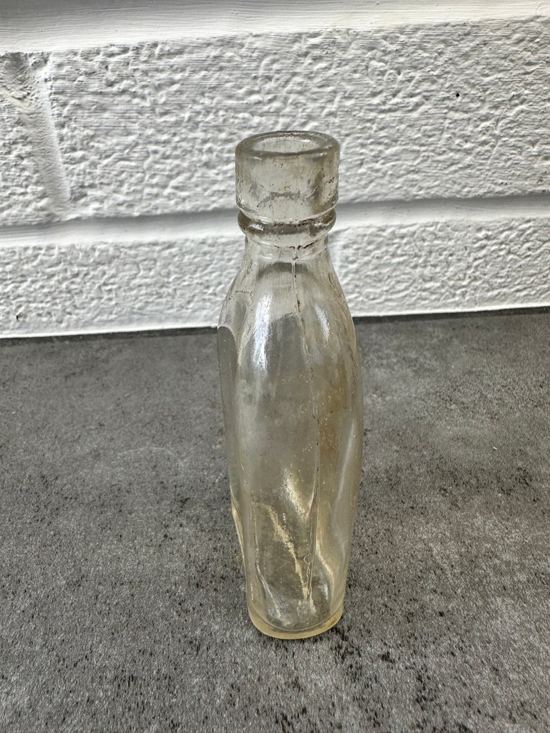 May include: A clear glass bottle with a rounded bottom and a slightly flared top. The bottle is empty and has a textured surface.
