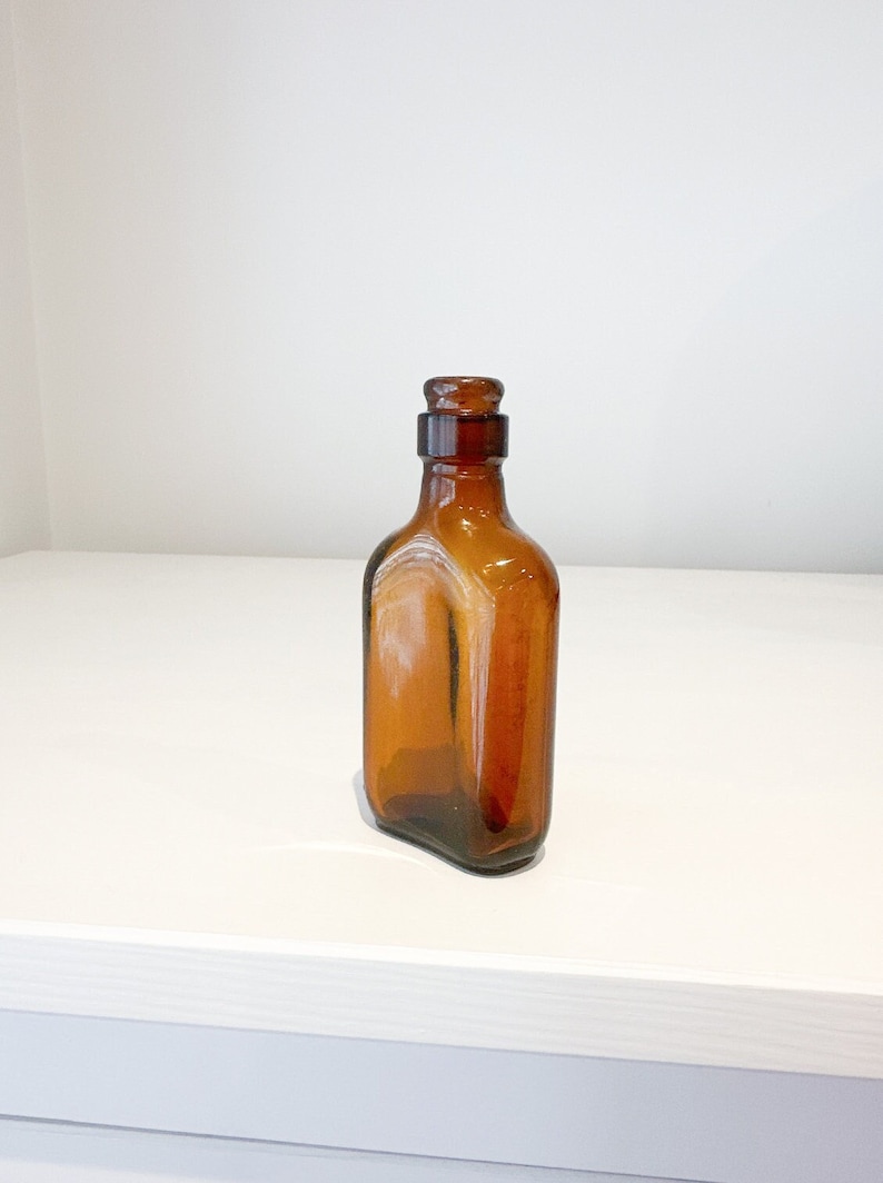 May include: A small, amber-colored glass bottle with a glass stopper. The bottle is empty and has a rectangular shape.