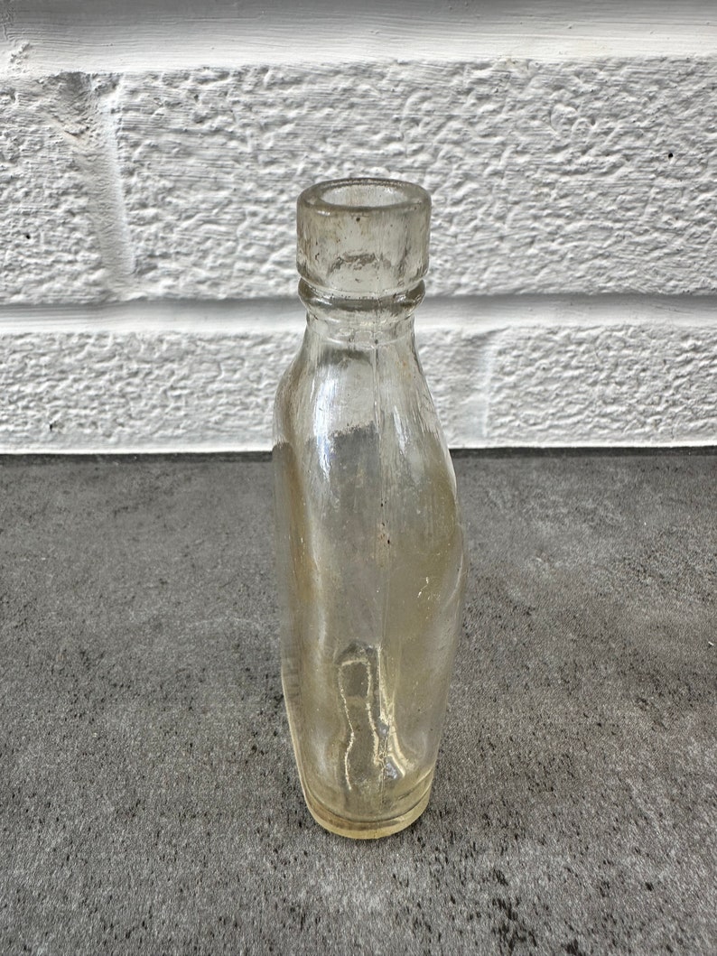 May include: A clear glass bottle with a rounded bottom and a slightly flared neck. The bottle is empty and appears to be vintage.
