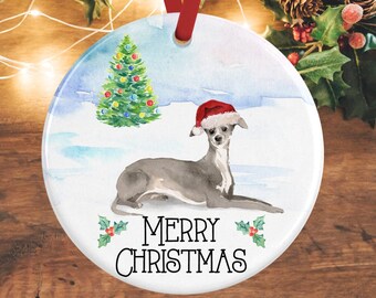 Può includere: Un ornamento rotondo in ceramica bianca con un'illustrazione ad acquerello di un levriero italiano grigio che indossa un cappello da Babbo Natale rosso. Sullo sfondo c'è un albero di Natale verde e la scritta MERRY CHRISTMAS in nero con agrifoglio.