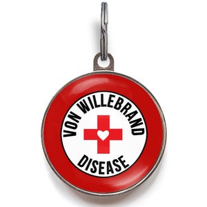May include: A round, red and white pet tag with the words "VON WILLBRAND DISEASE" encircling a red cross with a white heart in the center. The tag has a silver-colored metal loop at the top for attaching to a collar.