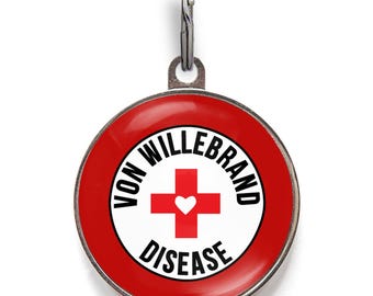 May include: A round metal pet identification tag with a red border and a white center. The center features a red cross with a small white heart in the middle. The text VON WILLEBRAND DISEASE is printed in black capital letters around the cross.
