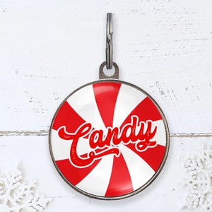 Può includere: Un medaglietta rotonda per animali domestici con un design a caramella alla menta piperita rossa e bianca. La parola "Candy" è scritta in rosso. La medaglietta ha un anello di metallo argentato.