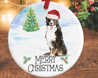 May include: A round ceramic ornament featuring a watercolour illustration of a Bernese Mountain Dog wearing a red Santa hat. The dog sits in a snowy landscape next to a decorated Christmas tree. The text MERRY CHRISTMAS is printed in black.