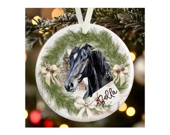 May include: A round white ceramic ornament featuring a black horse head centered within a green pine wreath. The wreath is decorated with cream-colored bows and pinecones. A small rectangular tag attached to the wreath displays the name Bella.