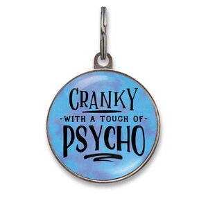 Könnte beinhalten: Eine runde Haustiermarke mit blauem Tie-Dye-Hintergrund und schwarzem Text, der CRANKY WITH A TOUCH OF PSYCHO lautet. Die Marke hat einen silberfarbenen Metallrand und einen kleinen Metallring an der Oberseite zur Befestigung am Halsband.