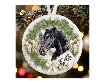 May include: A round white ceramic ornament featuring a black horse head centered within a green pine wreath. The design includes three cream-colored bows, pinecones, and a small rectangular tag with the name Bella written in black script.